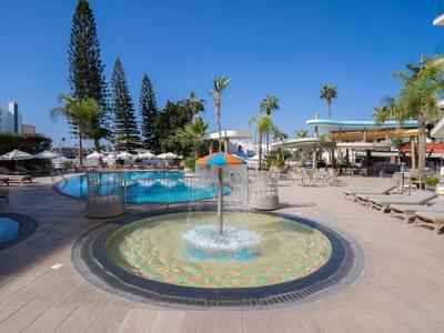 Moderner Hotelpoolbereich mit Springbrunnen, Liegestühlen und mediterraner Vegetation unter klarem Himmel.