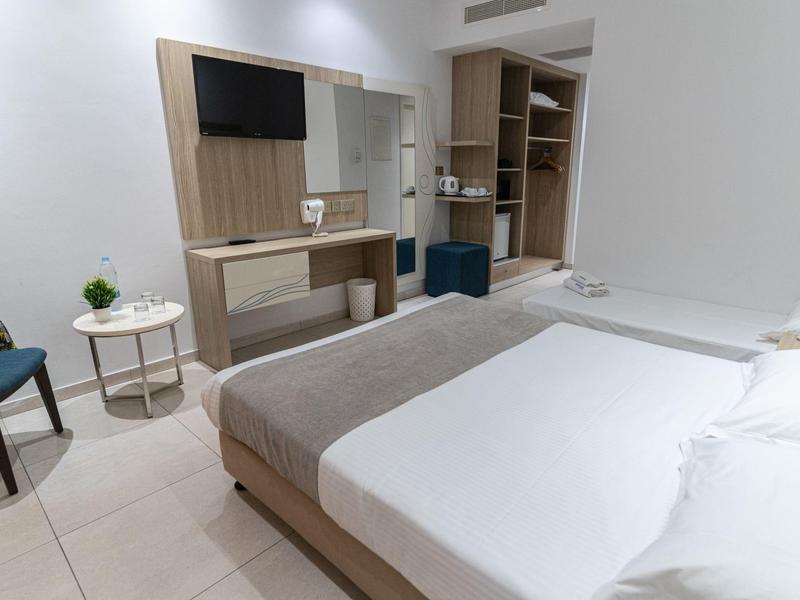 Modern hotel room with double bed, TV, desk, and open wardrobe in neutral tones.