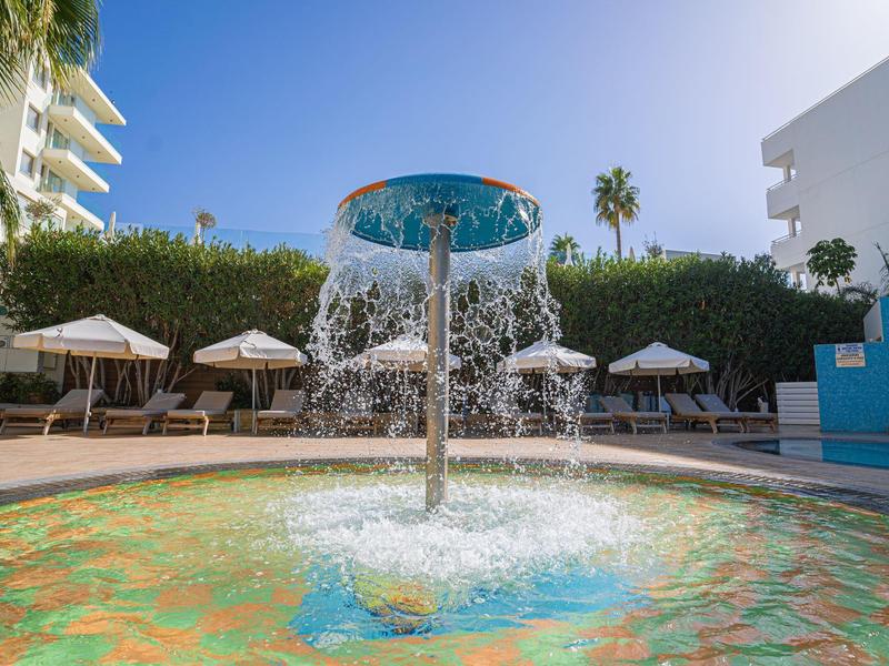 Fountain with clear water surrounded by sun umbrellas and palm trees in a hotel garden.