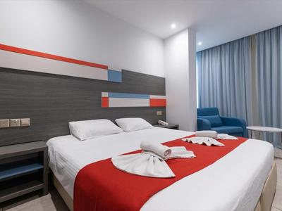 Modernes Hotelzimmer mit großem Bett, roten Akzenten, blauen Sesseln und hellen Vorhängen.