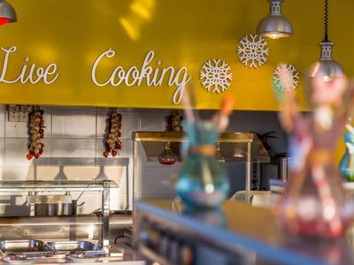 Küchenzeile mit Hängelampen, Gewürzen und Schriftzug "Live Cooking" auf gelber Wand.