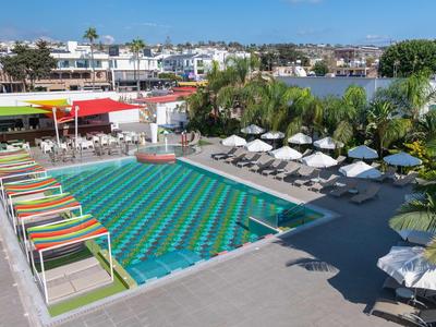 Außenpool mit bunten Liegestühlen und weißen Sonnenschirmen, umgeben von Palmen und Gebäuden.