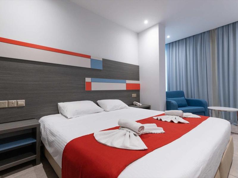 Modernes Hotelzimmer mit großem Bett, roten Akzenten, blauen Sesseln und hellen Vorhängen.