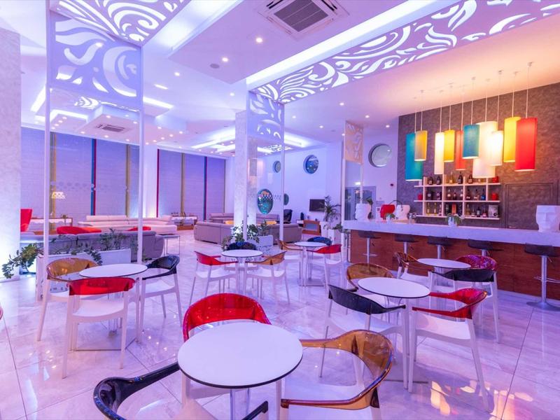 Moderner Café-Innenraum mit bunten Stühlen, weißen Tischen und bunter LED-Beleuchtung an Decke und Wand.