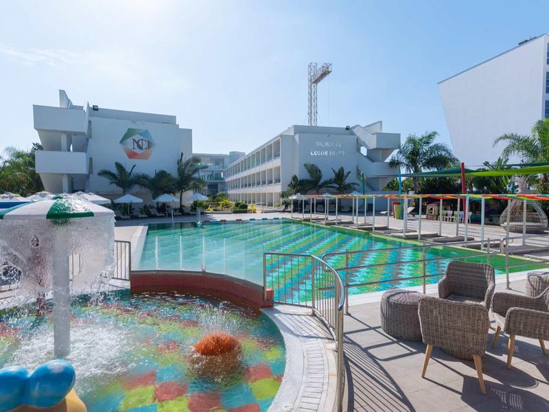 Moderner Außenpoolbereich mit Wasserspielen, Liegestühlen und Palmen unter blauem Himmel.