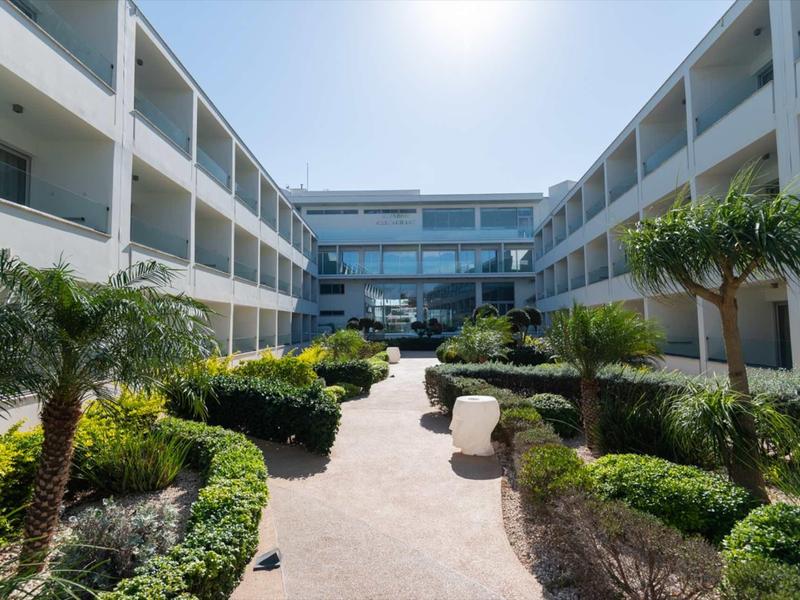 Moderner Gebäudekomplex mit zwei Stockwerken, begrünter Innenhof und palmähnlichen Pflanzen.