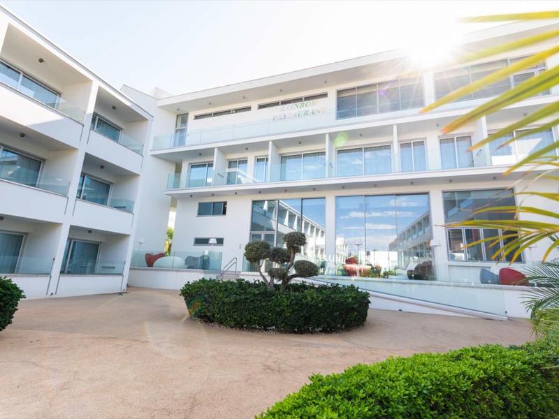 Modernes weißes Gebäude mit vielen Fenstern und begrüntem Innenhof bei Sonnenschein.