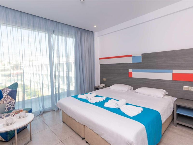 Modernes Hotelzimmer mit Doppelbett, blauem Bettläufer, großen Fenstern und Vorhängen.