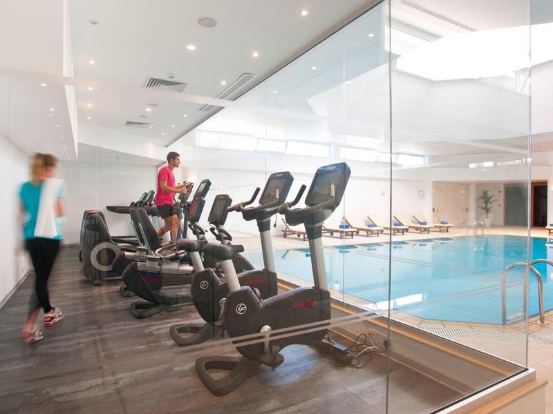 Modern gym equipment next to an indoor pool with glass wall in a hotel.