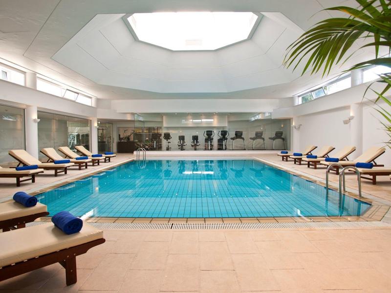 Bright indoor pool with loungers, plants, and fitness equipment in a modern setting.