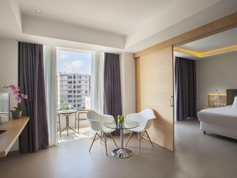 Modern hotel room with seating area, glass balcony, and view of surrounding buildings.