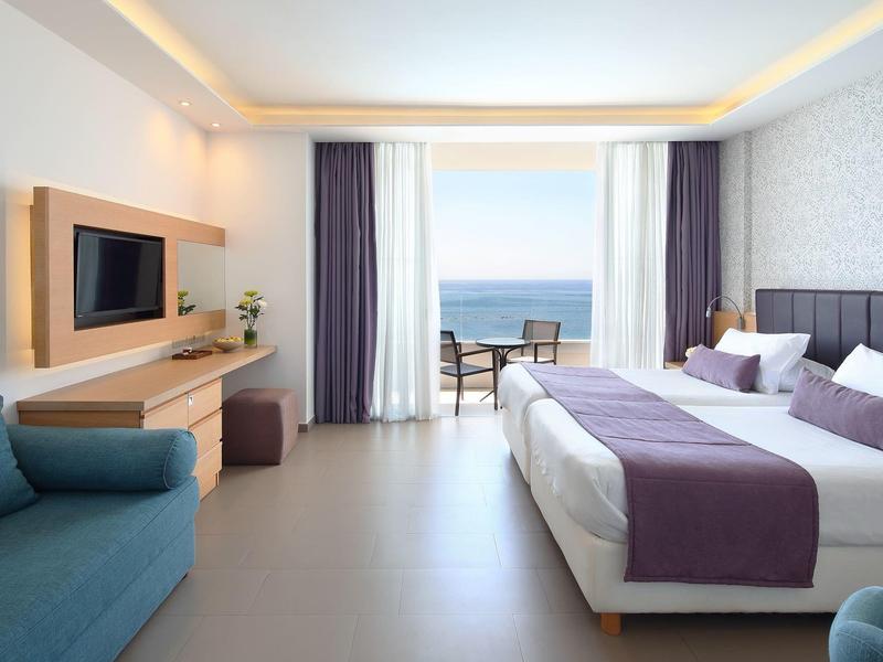 Modern hotel room with double bed, sofa, and sea view through large windows.