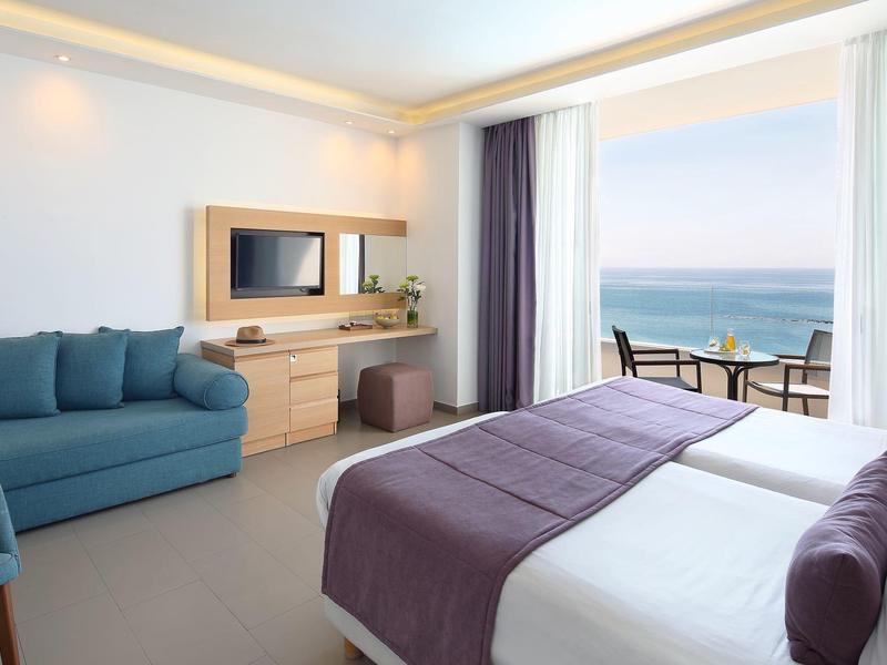 Bright hotel room with double bed, blue sofa, and balcony overlooking the sea.