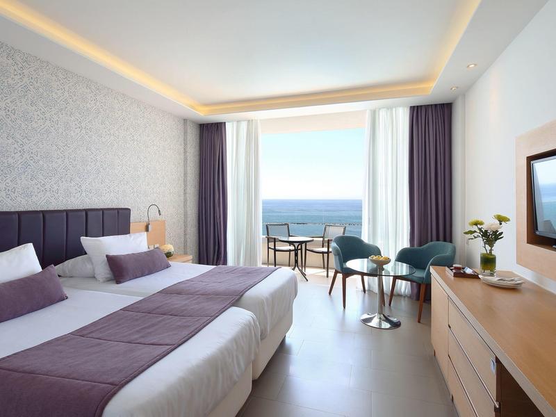 Modern hotel room with two beds, sea view, and balcony seating area.