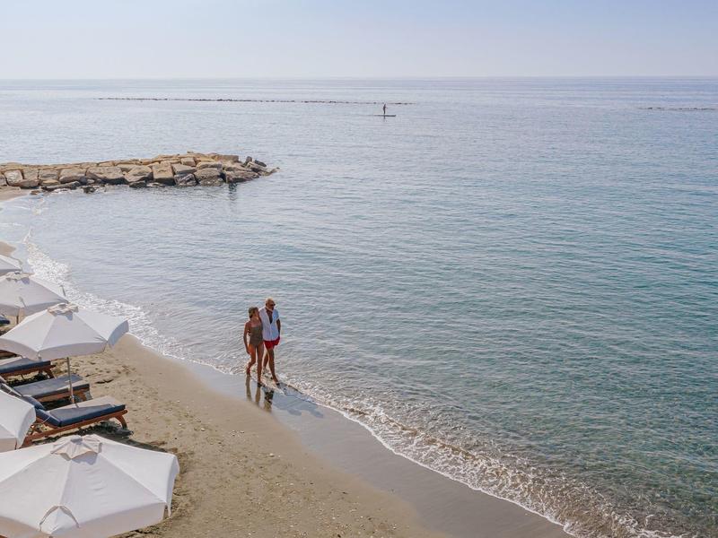 Couple walking on the beach with lounge chairs and umbrellas, facing calm sea.
