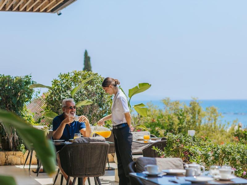 A man sits at an outdoor table with sea view, served by a waitress.
