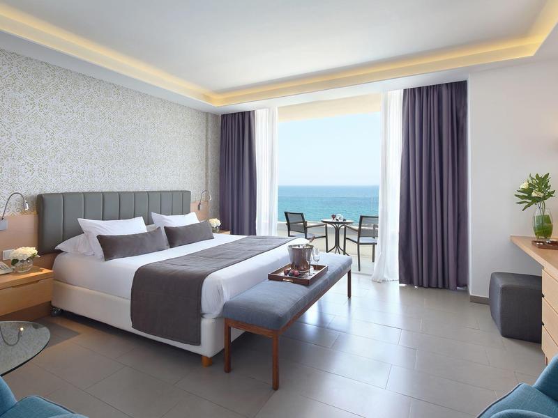 Bright hotel room with large bed, sea view, and balcony with chairs.