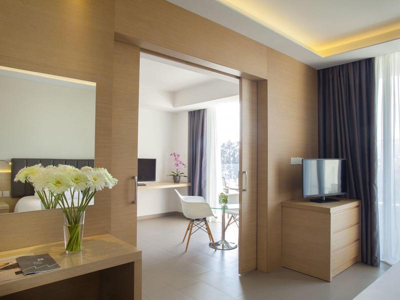 Bright hotel room with wooden dresser, TV, and open doorway to living area with balcony.