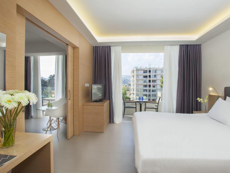 Modern hotel room with large bed, balcony overlooking buildings, and fresh flowers.