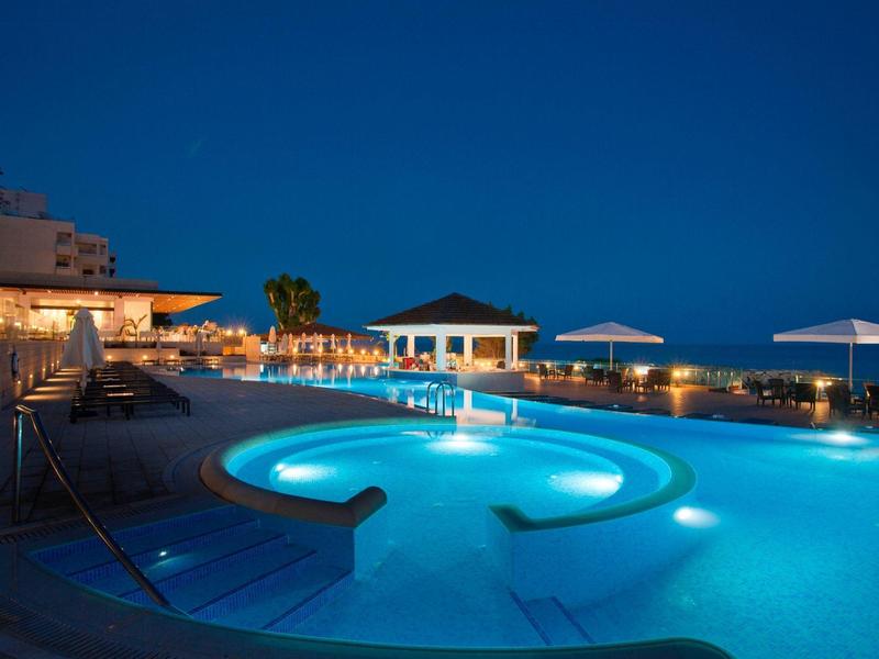 Illuminated hotel pool at night with seating area and sea view.
