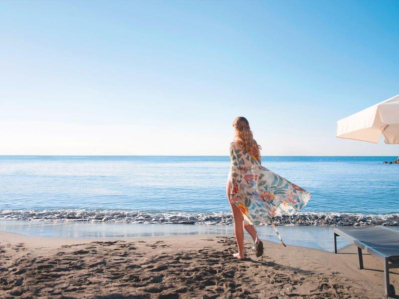 Woman on beach beside lounge chair and umbrella overlooking the sea.