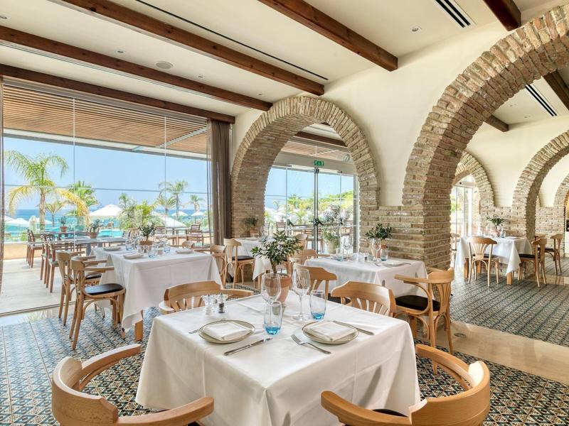 Elegant restaurant with white tablecloths, wooden chairs, and large windows overlooking palm trees.