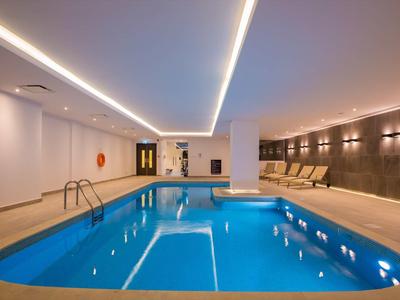 Piscina coperta moderna con piastrelle blu, lettini e illuminazione indiretta.