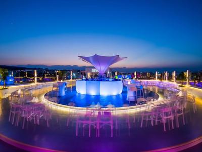 Moderne Terrasse mit runden Tischen, bunten Lichtern und zentraler blauer Skulptur bei Abenddämmerung.