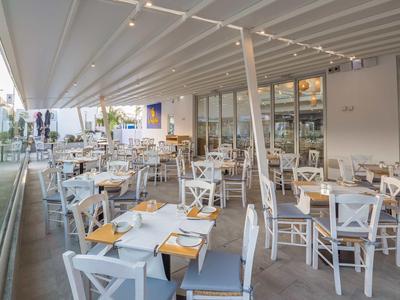 Area esterna moderna di un ristorante con sedie bianche e tavoli in legno accanto a una facciata in vetro.