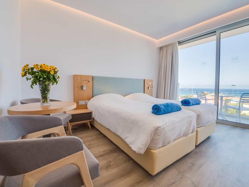 Helles Schlafzimmer mit Doppelbett, zwei blauen Handtüchern, Blumen auf dem Tisch und Meerblick durch Glasfenster.