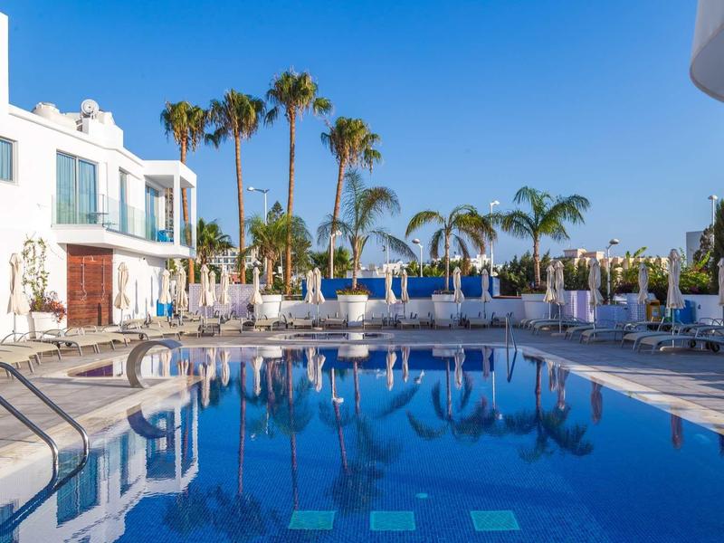 Großer blauer Pool neben weißem Hotel mit Palmen und Liegestühlen unter klarem Himmel.