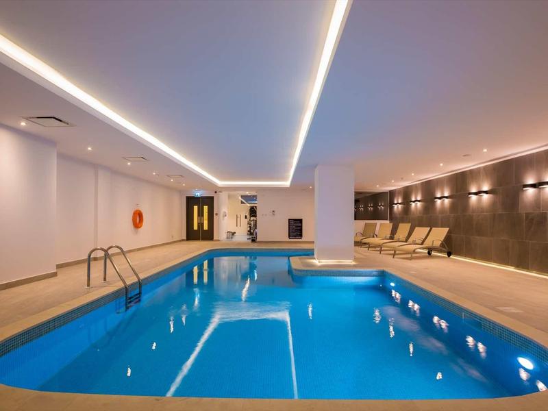 Innenraum mit rechteckigem Hallenbad, weißen Wänden, LED-Deckenbeleuchtung und beigen Liegestühlen.