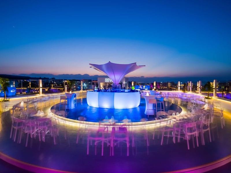 Moderne Terrasse mit runden Tischen, bunten Lichtern und zentraler blauer Skulptur bei Abenddämmerung.