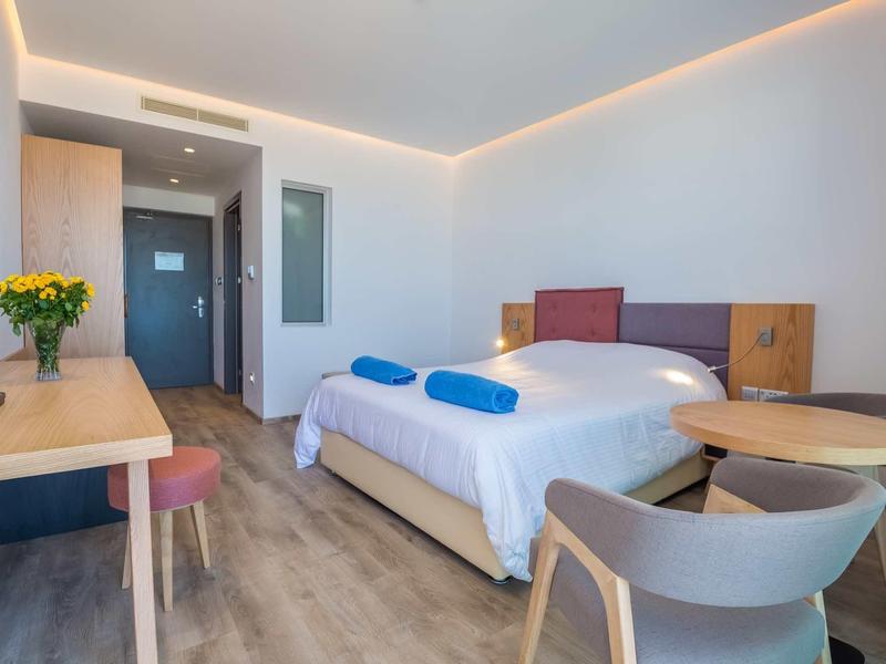 Helles Hotelzimmer mit Doppelbett, Holzmöbeln, runden Tisch, Stühlen und indirekter Deckenbeleuchtung.