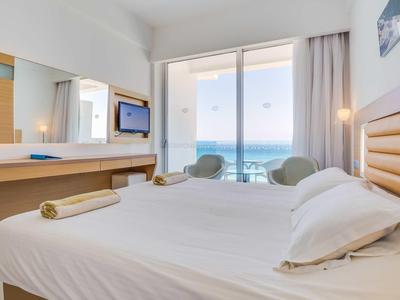 Jasny pokój hotelowy z podwójnym łóżkiem, dwoma fotelami i widokiem na morze przez duże okna.