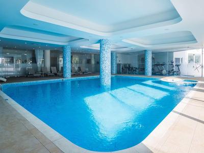 Jasny basen kryty z dużymi oknami, niebieską wodą i białymi kolumnami.