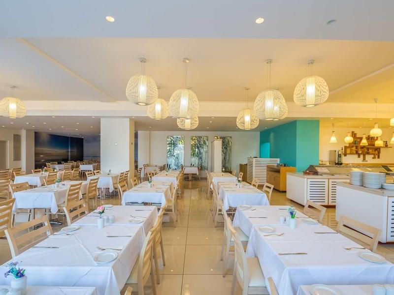 Jasna, nowoczesna restauracja z białymi stołami, drewnianymi krzesłami i ciepłym oświetleniem sufitowym.