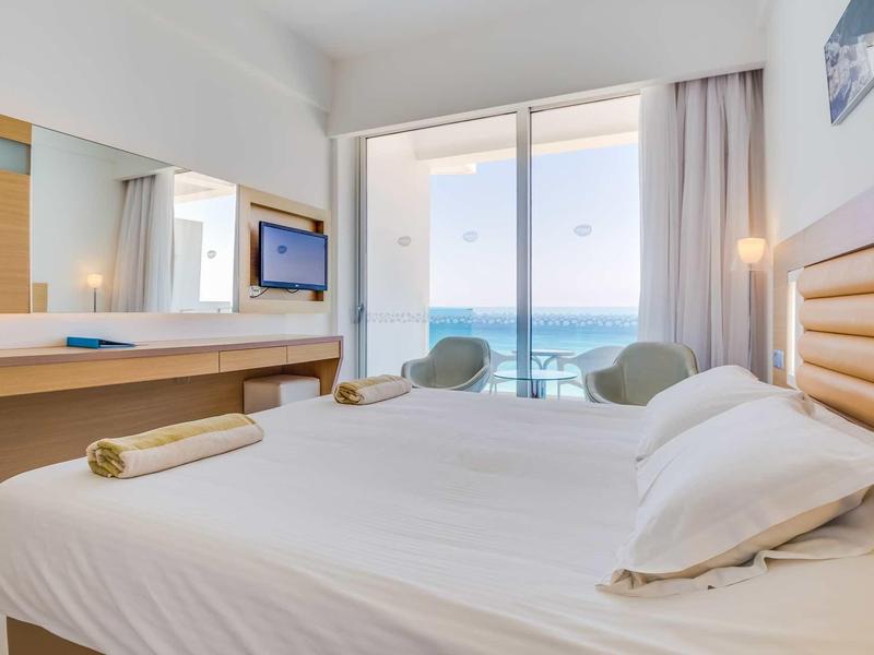 Jasny pokój hotelowy z podwójnym łóżkiem, dwoma fotelami i widokiem na morze przez duże okna.
