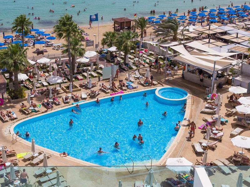 Zatłoczony basen hotelowy z parasolami i plażą blisko morza pod jasnym niebem.