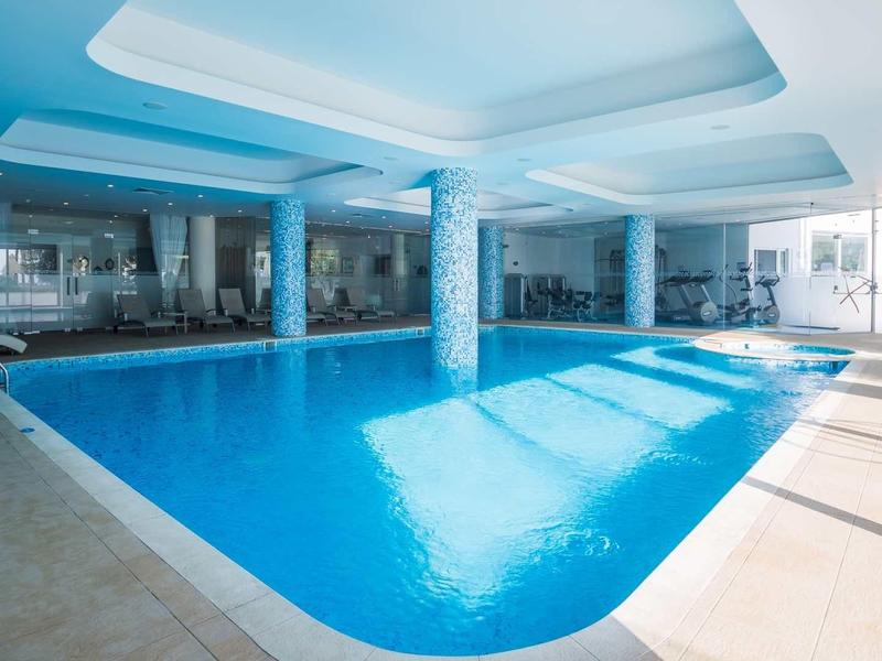 Jasny basen kryty z dużymi oknami, niebieską wodą i białymi kolumnami.