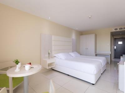 Modern hotel room with white bed, small table, and chairs on a light floor.