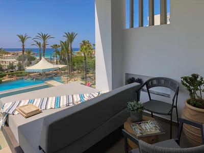 Balcony with seating, plants, pool view in a hotel with sea view and palm trees.