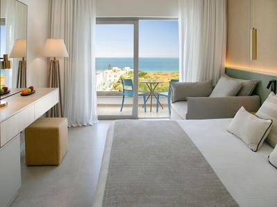 Helles Hotelzimmer mit großem Fenster, Meerblick und Terrasse mit Tisch und Stühlen.