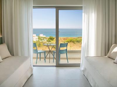 Blick aus einem hellen Zimmer auf eine Terrasse mit Stühlen und Meer im Hintergrund.