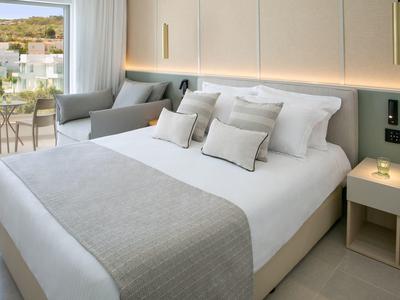 Modernes Hotelzimmer mit großem Bett, beigen und grauen Akzenten und Balkon mit Blick auf grüne Hügel.