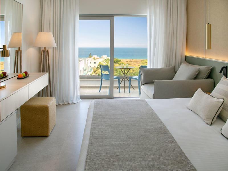 Modernes Hotelzimmer mit Meerblick, Doppelbett, Sofa und Zugang zu einem Balkon mit Tisch und Stühlen.