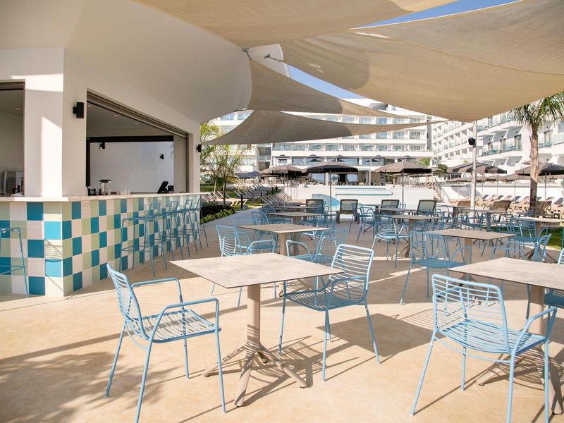 Außenbereich einer Hotelbar mit blauen Stühlen und Tischen, Sonnensegeln und Hotelgebäude im Hintergrund.