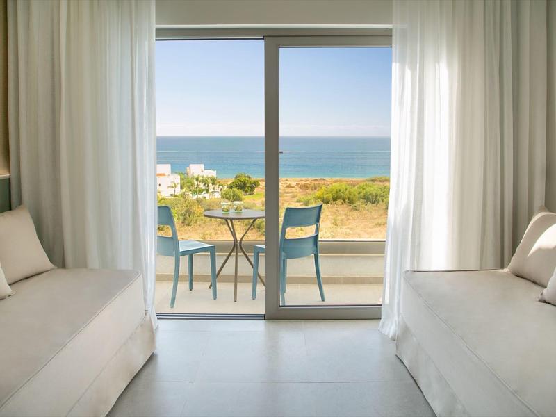 Blick aus einem hellen Zimmer auf eine Terrasse mit Stühlen und Meer im Hintergrund.