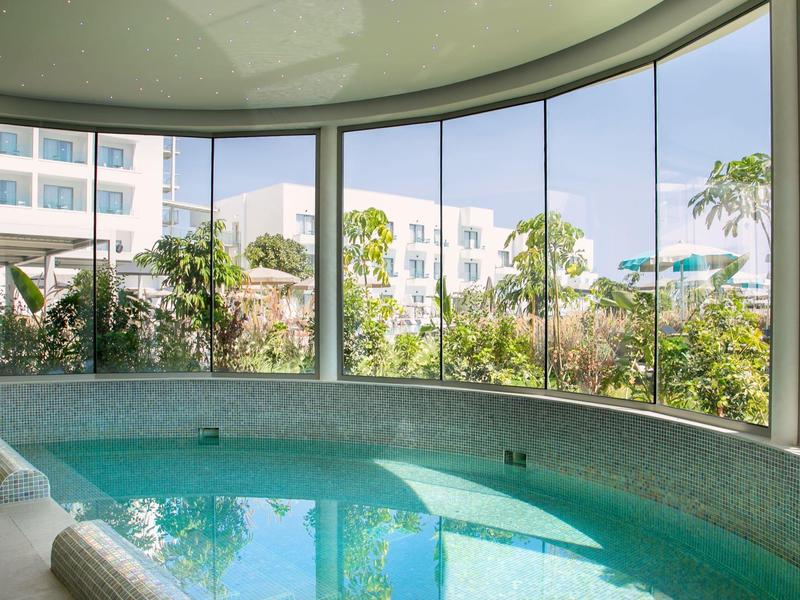 Runder Innenpool mit Panoramafenstern und Blick auf Hotel und Palmen.