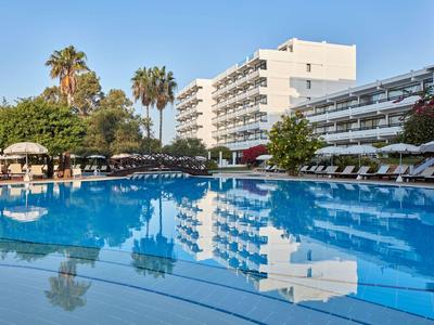 Großer Hotelpool mit Liegen und Palmen vor einem mehrstöckigen Gebäude bei klarem Himmel.